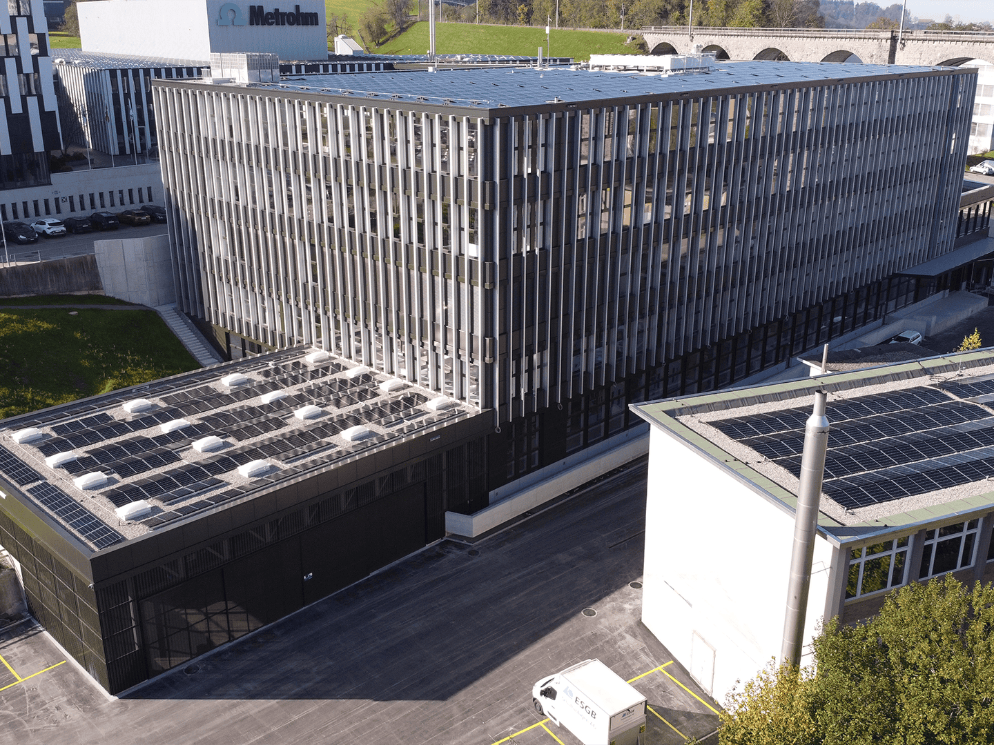 Aerial view of a modern office building with solar panels on the roof, surrounded by parking spaces and greenery, showcasing sustainable architecture and renewable energy integration.