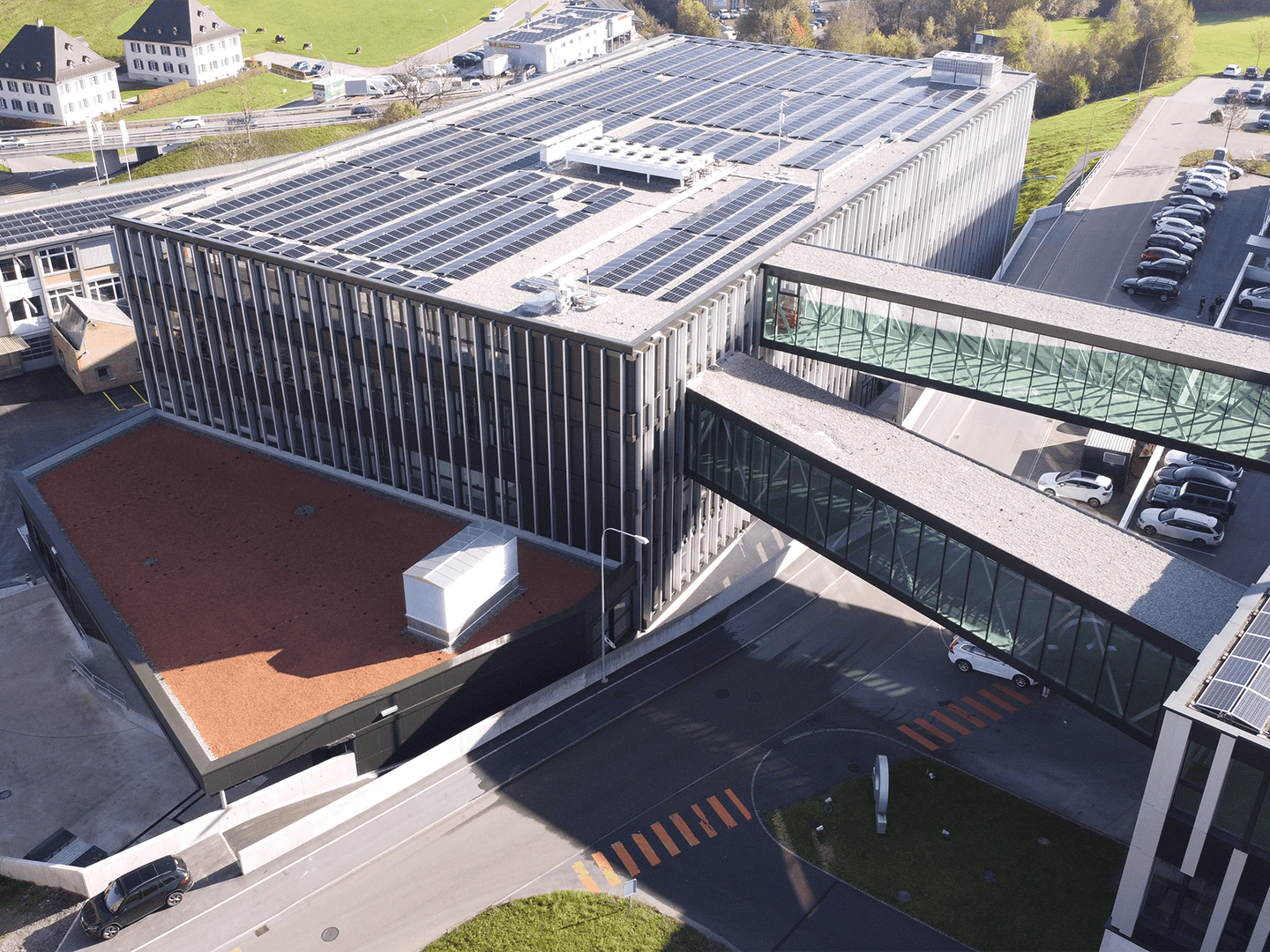 Solar panels installed on the roof of a modern office building with glass walkways connecting different sections, parking lot with cars, and surrounding green landscape.