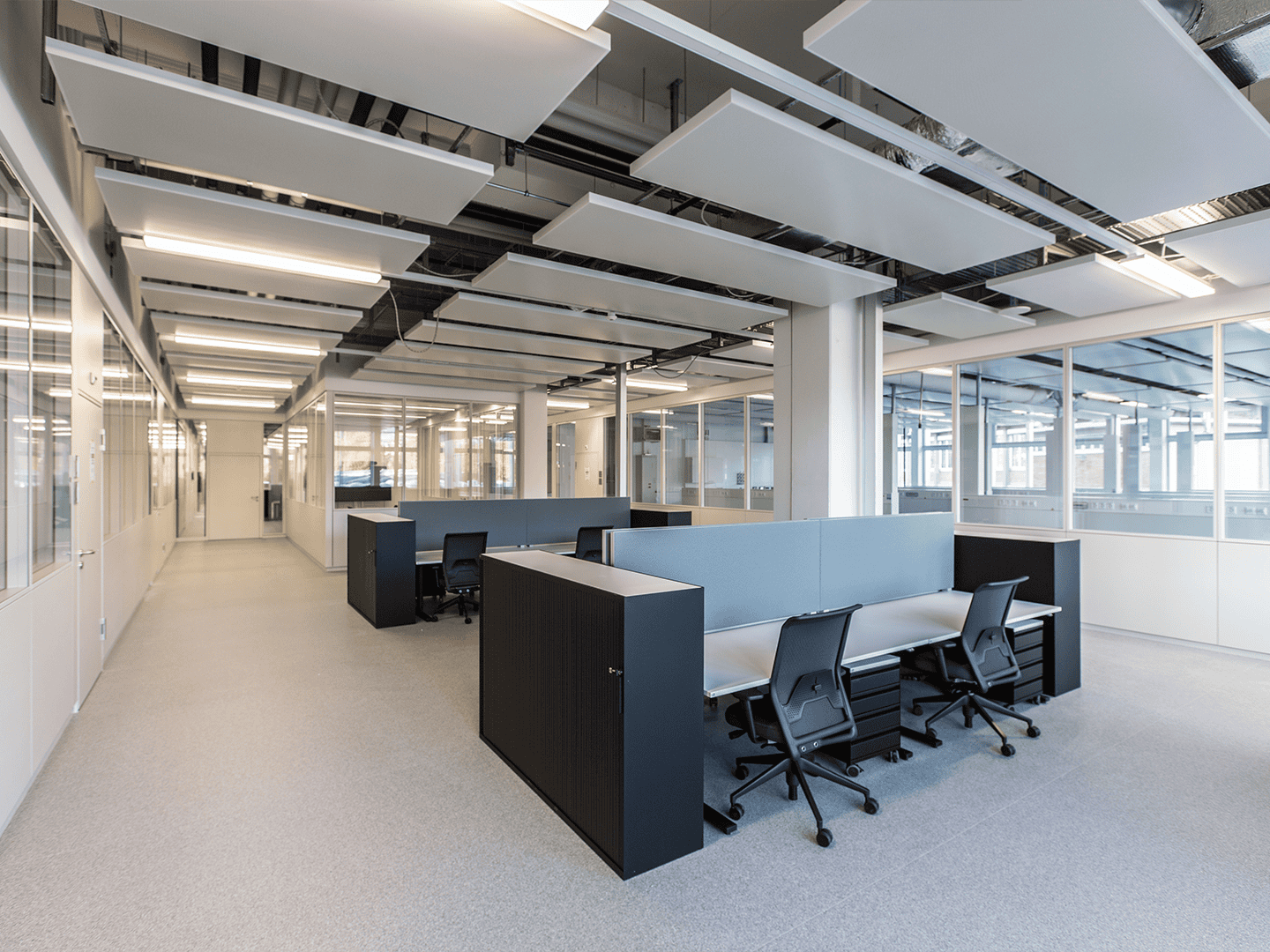 Office workspace with modern cubicles, black chairs, and glass-walled meeting rooms illuminated by ceiling lights and natural daylight.