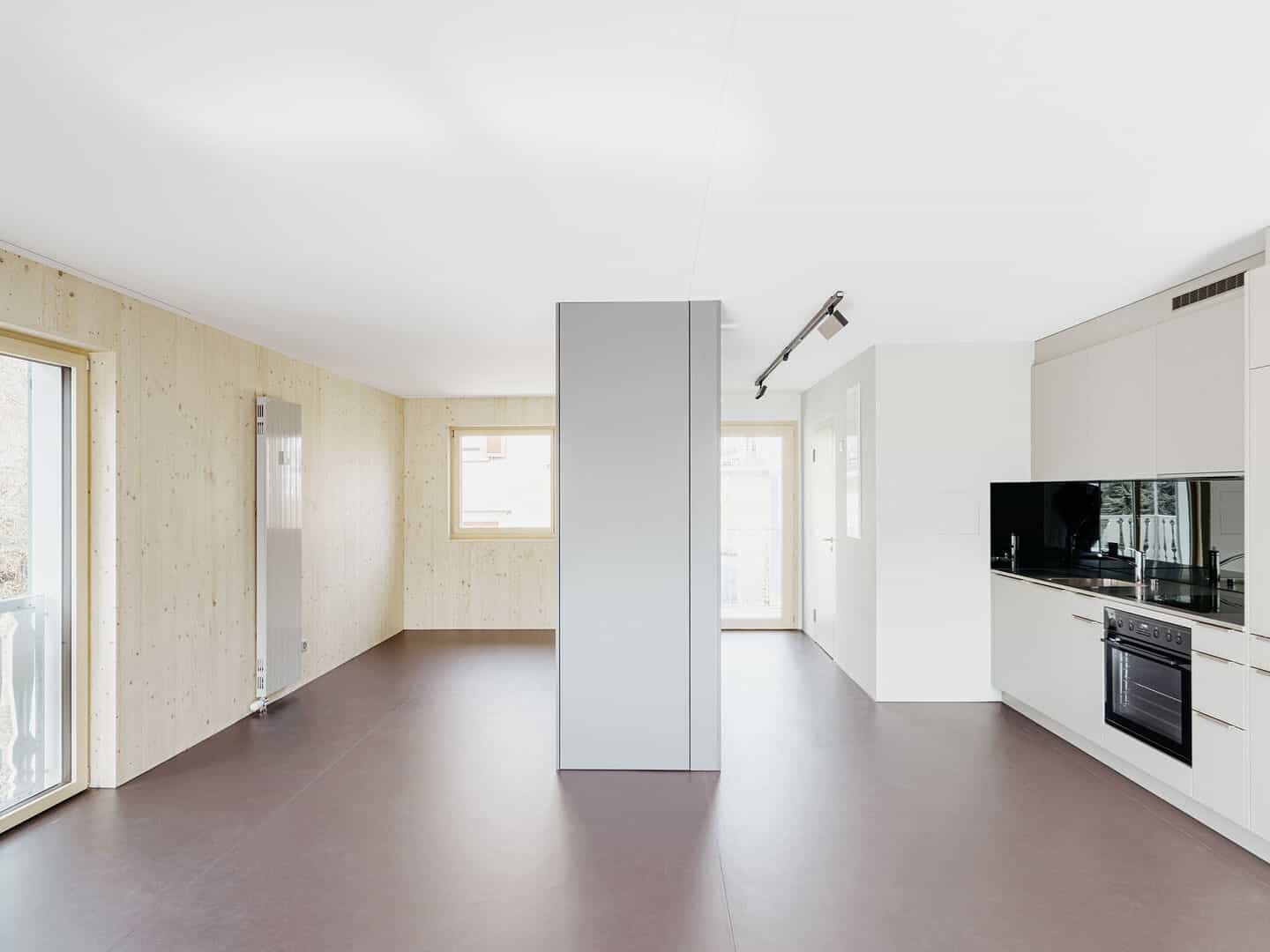 Modern minimalist kitchen and living space with white cabinetry, black appliances, and large windows providing natural light in an open floor plan.