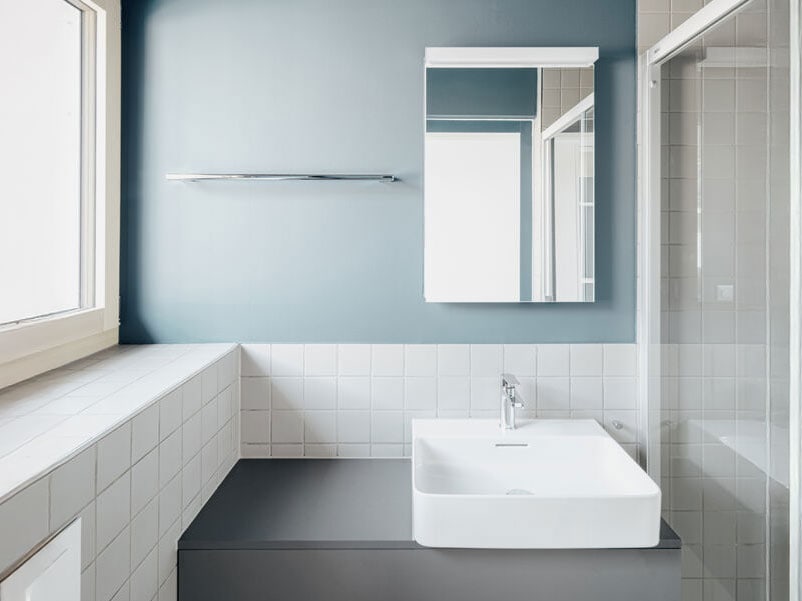 Modern bathroom with a white sink, mirror, and shower enclosure, featuring a minimalist design with blue and white tiles and natural light from a large window.