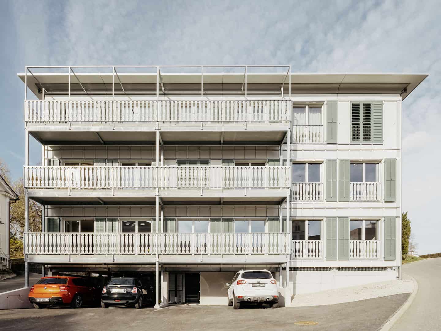 Modern multi-story residential building with open balconies, green shutters, and an underground parking area with several cars. The structure features a clean, minimalist design with white and light gray elements.