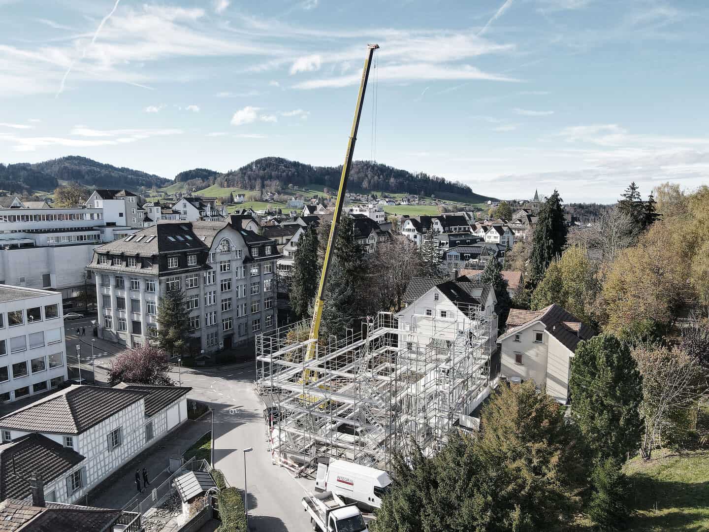 Heavy construction crane lifting materials at a building site amidst scaffolding in a suburban area with residential and commercial buildings, surrounded by lush trees and rolling hills.