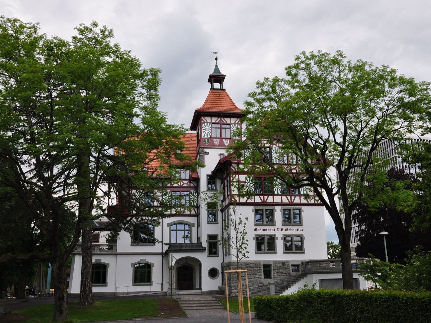 Colorful historic building with a red-tiled roof, white walls, and exposed wooden beams, partially obscured by lush green trees, featuring a small tower with a weather vane, set in an urban environment.