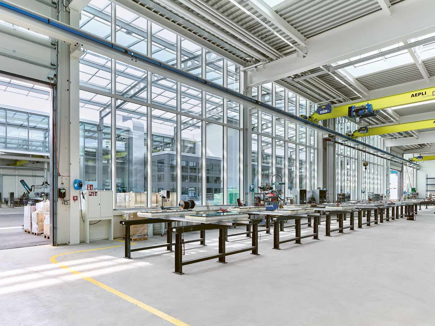 Factory interior with large glass windows, workbenches, and overhead cranes in an industrial manufacturing facility.
