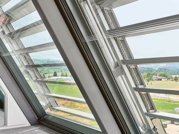 - A close-up view of a modern, tilted window with adjustable metal blinds, overlooking a green landscape with a distant house and trees, under a bright sky.