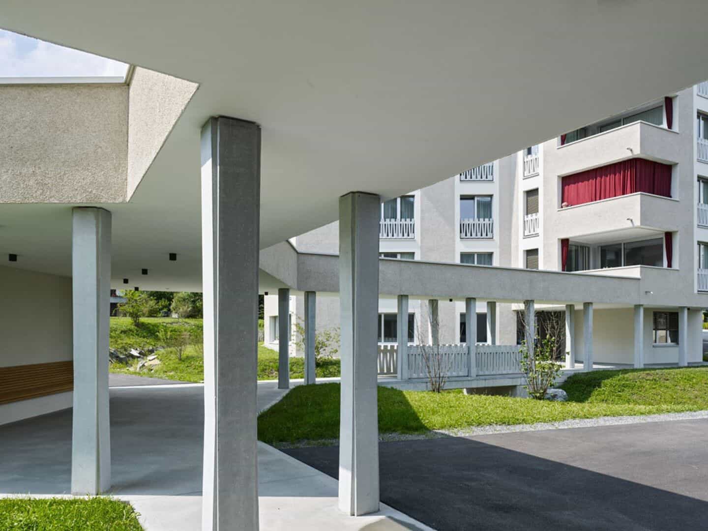 Modern residential apartment building with balconies, white exterior, and red curtains, featuring a courtyard with greenery and covered walkways.