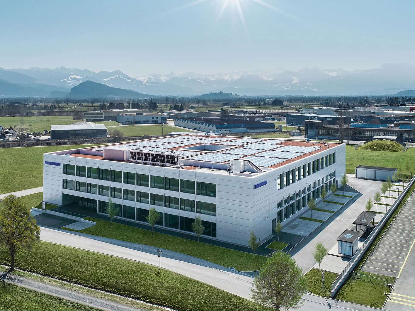 Modern office building with solar panels on the roof, surrounded by green lawns and parking lot, with mountains in the background and clear blue sky.