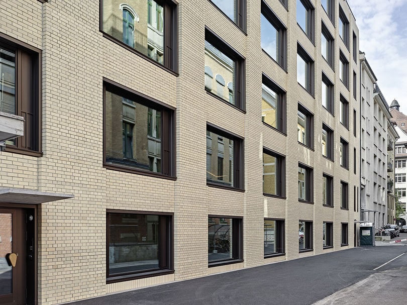Modern beige brick apartment building with large dark-framed windows on a city street. The building features multiple floors and a contemporary architectural design with clean lines.