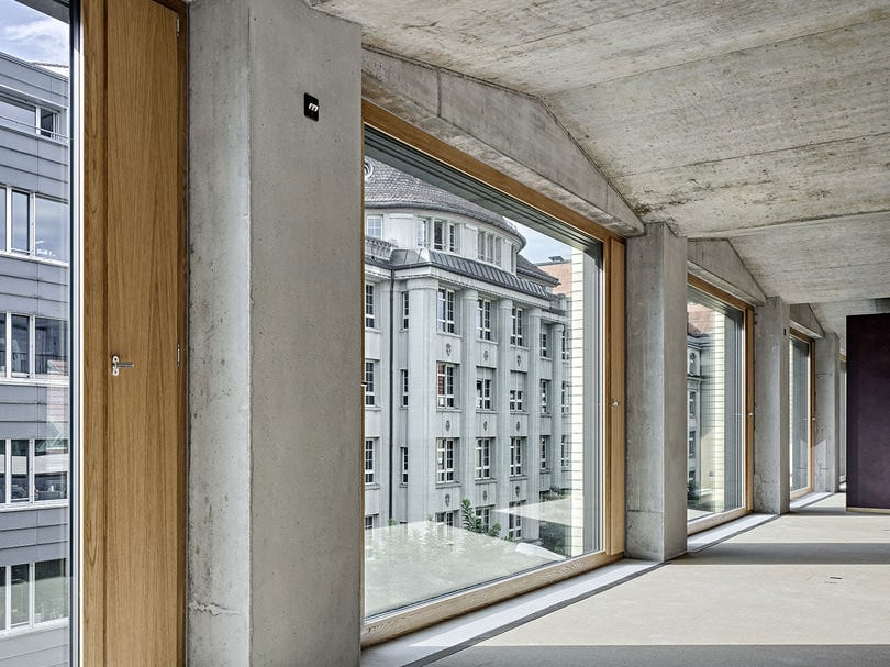 Floor-to-ceiling windows with concrete and wooden framing in a modern building, overlooking a traditional multi-story city building, interior view.