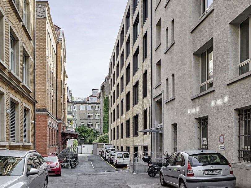 A narrow urban alleyway between modern and older multi-story buildings, with parked cars and scooters, leading to a slightly elevated area with additional structures and greenery in the background.