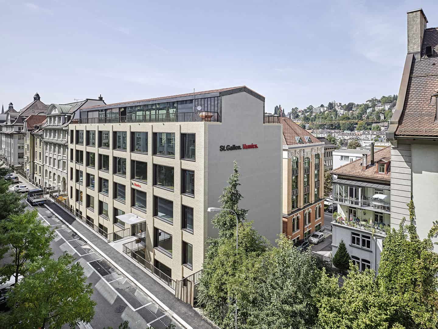 Modern multi-story building with large windows and a rooftop terrace in an urban neighborhood, surrounded by older residential buildings and greenery.
