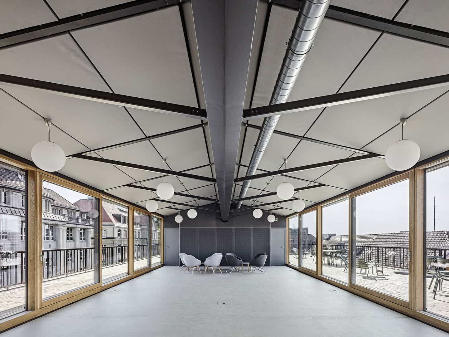 Modern rooftop pavilion with large glass windows, stylish seating area, and unique ceiling design featuring black geometric structures and round white pendant lights.