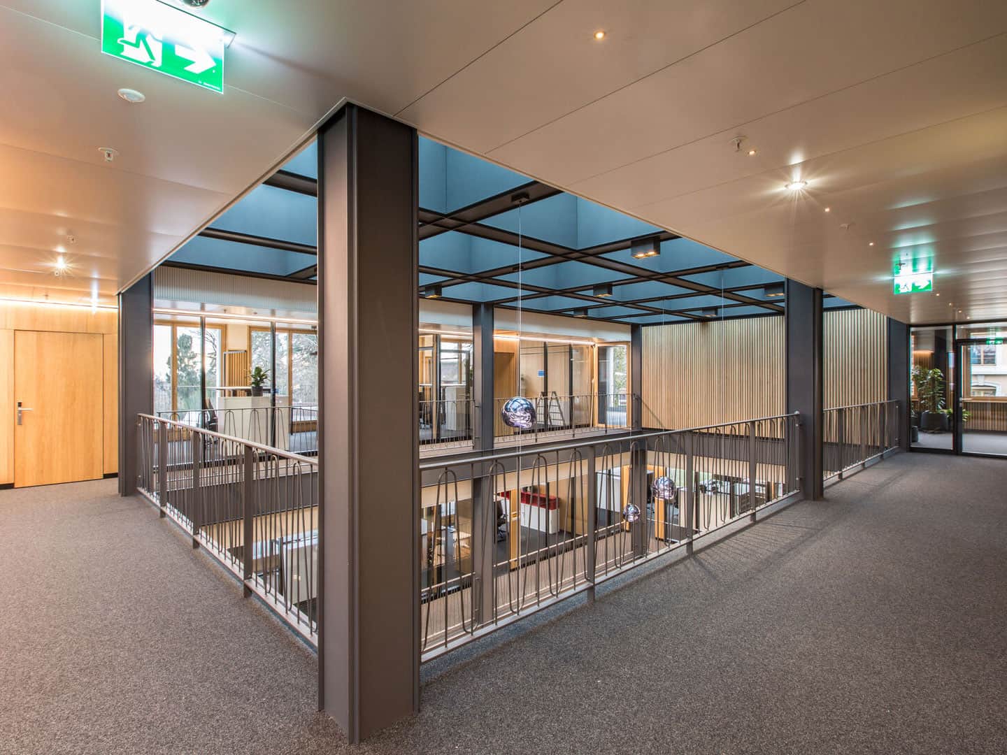 Modern indoor office hallway with glass walls, wooden accents, and metal railings overlooking a lower level, illuminated by natural light and ceiling lighting, creating a bright, open workspace environment.