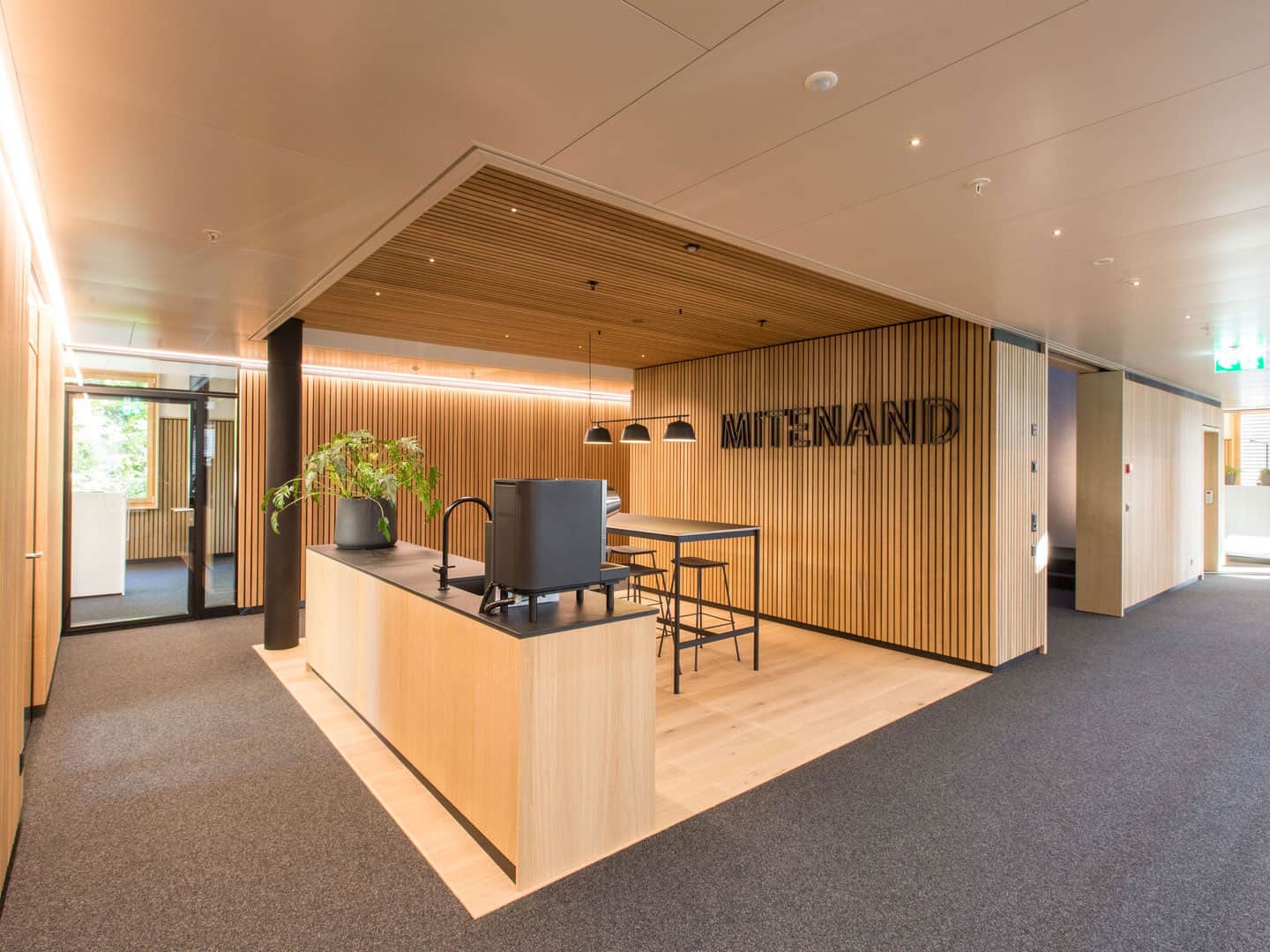 Modern office reception area with wood paneling, a reception desk with a black machine, potted plant, and the sign "MITENAND" on the wall. Bright, welcoming space with natural light from windows.