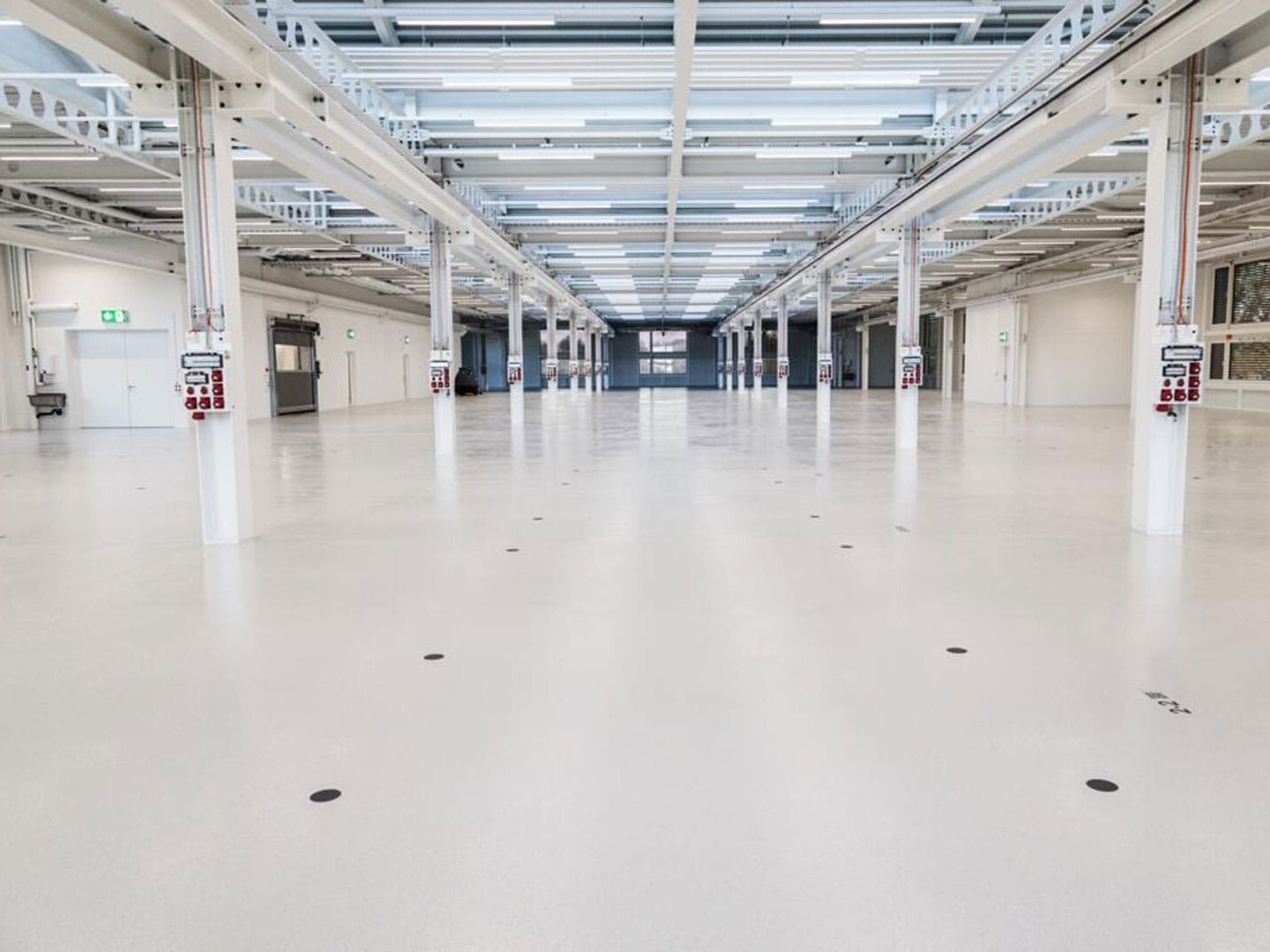 Empty industrial warehouse with white polished concrete floors, exposed ceiling pipes, and structural columns. Well-lit with natural and artificial lighting, ready for storage or manufacturing setup.