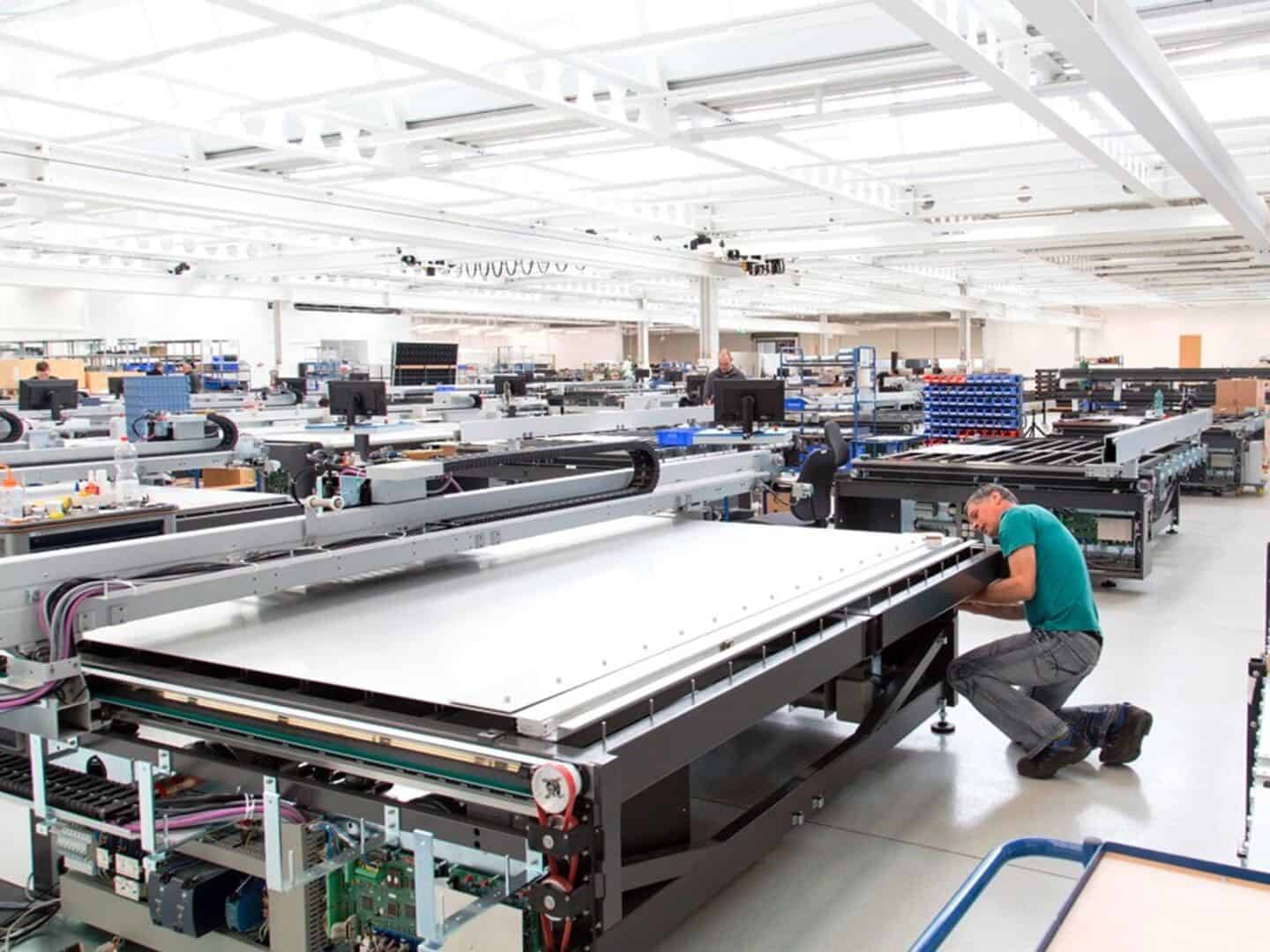 Advanced manufacturing cleanroom with technicians assembling large flat-panel displays or electronic components in a spacious, well-lit factory environment.