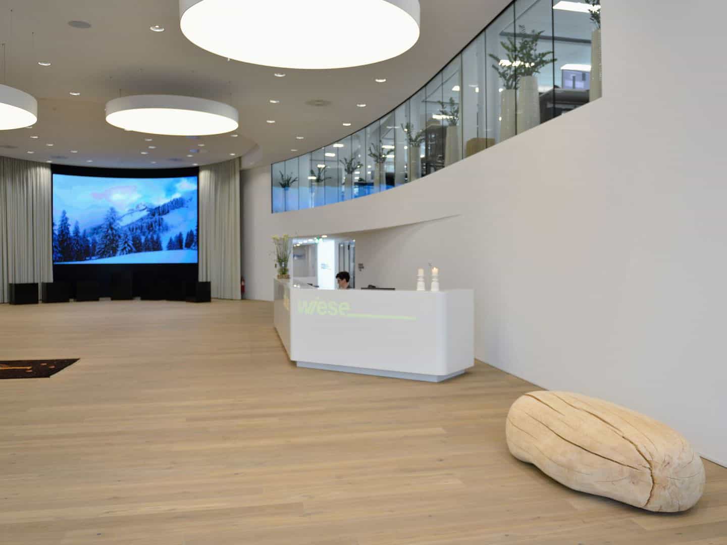 Bright modern hotel lobby with a large digital mountain landscape screen, white reception desk, and stylish decor including a large beige stone sculpture on the light wood floor.