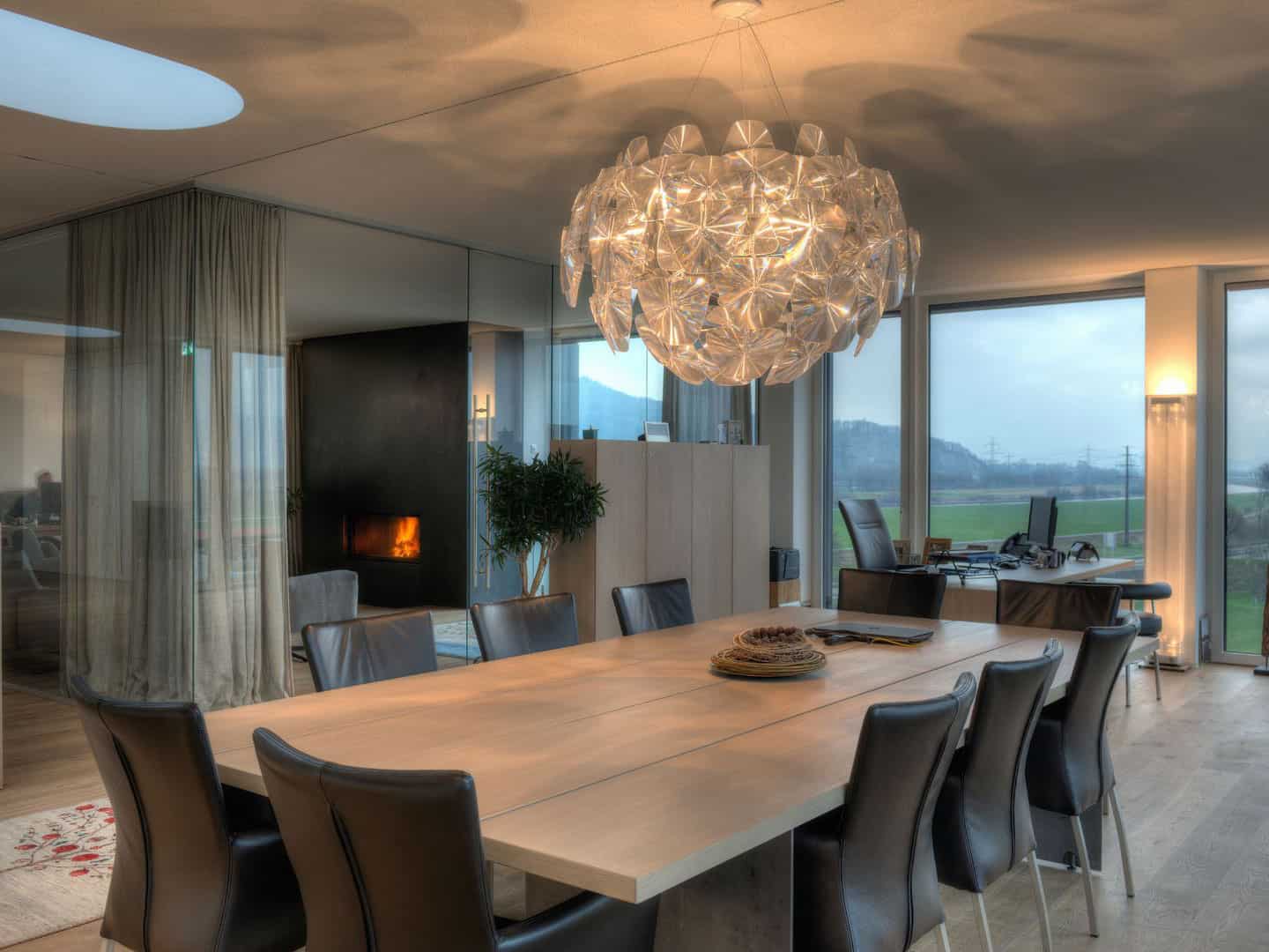 Elegant modern dining room with a large wooden table, black leather chairs, and a striking crystal chandelier lighting fixture. The space features floor-to-ceiling windows with a scenic view and a cozy fireplace.