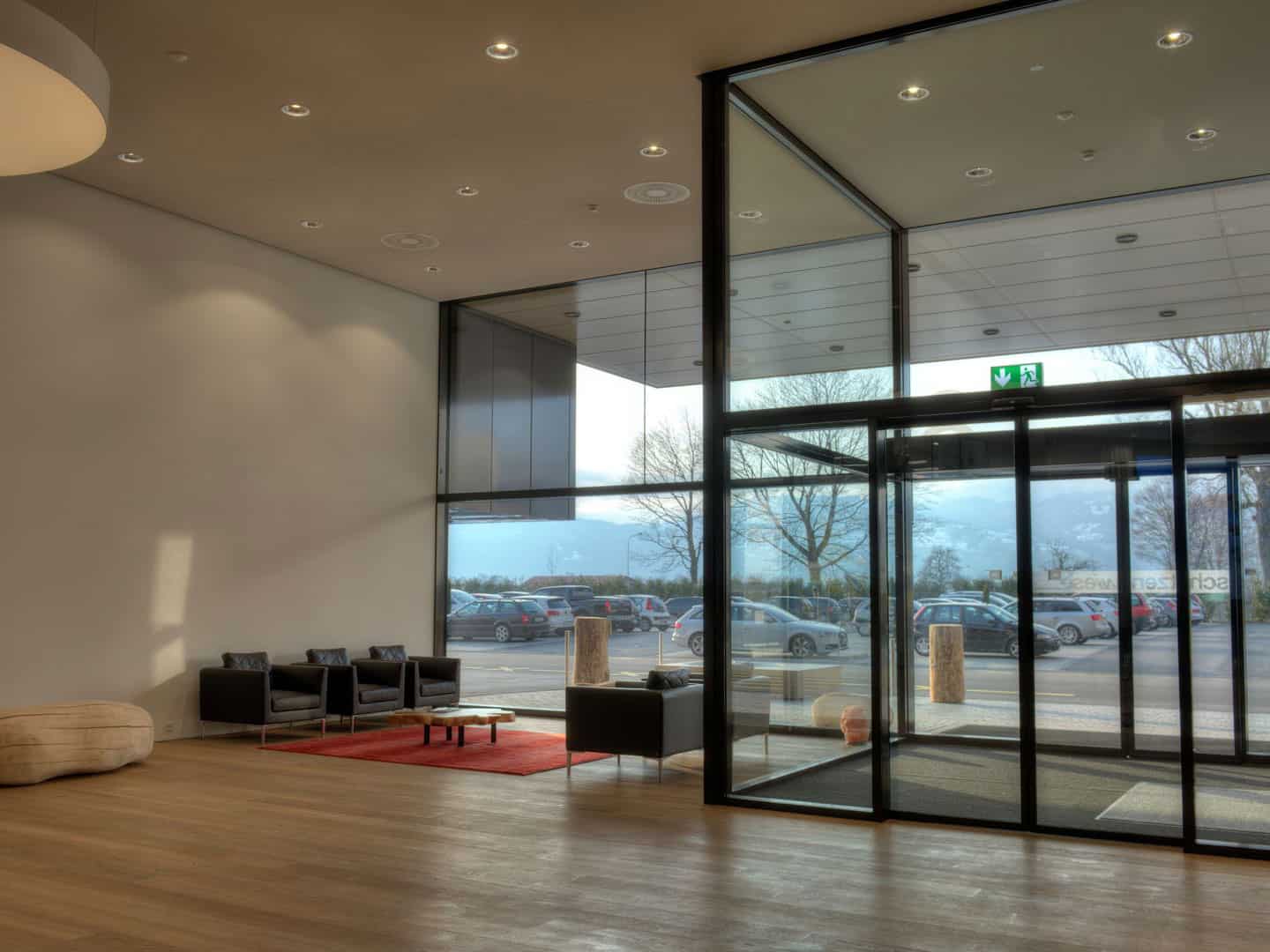 2. Modern hotel lobby with minimalist design, large glass entrance doors, wooden flooring, and seating area with black chairs and a red rug.