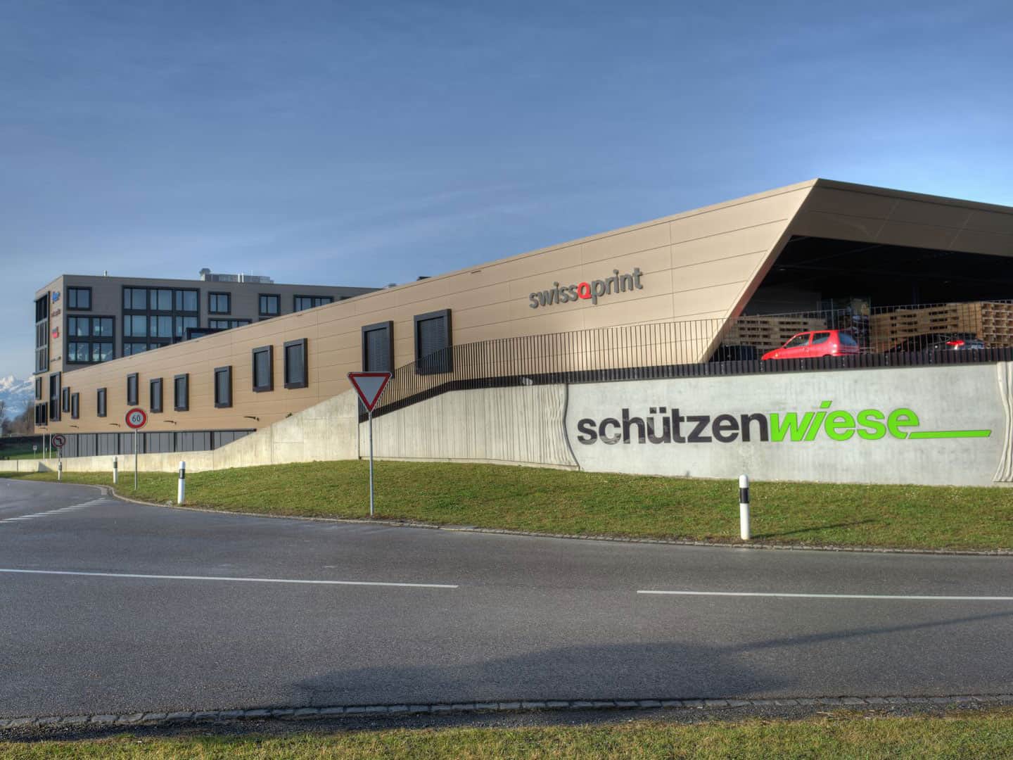 Modern commercial building with the signage "swissQprint" and "schützenwiese" under a clear blue sky, featuring a parking garage with red vehicles and a wooden fence. The structure has angular design elements and large windows.