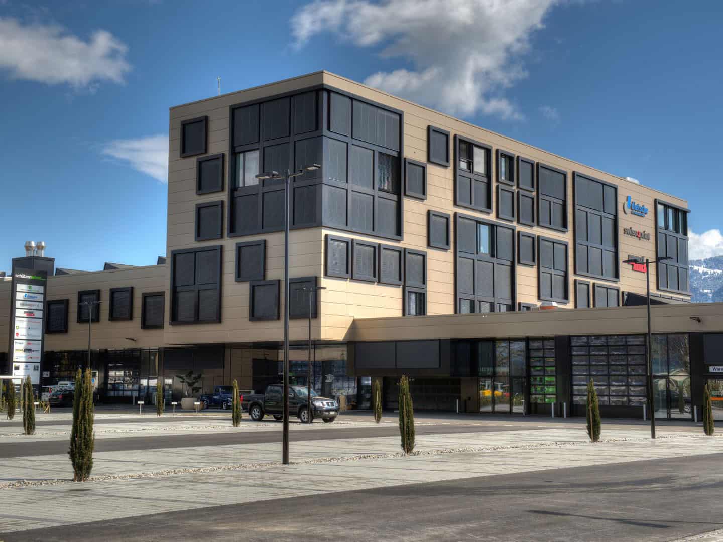 Modern multi-story commercial building with large glass windows and signage, situated in a cityscape with a clear blue sky, surrounded by a paved area with small trees and parked cars.
