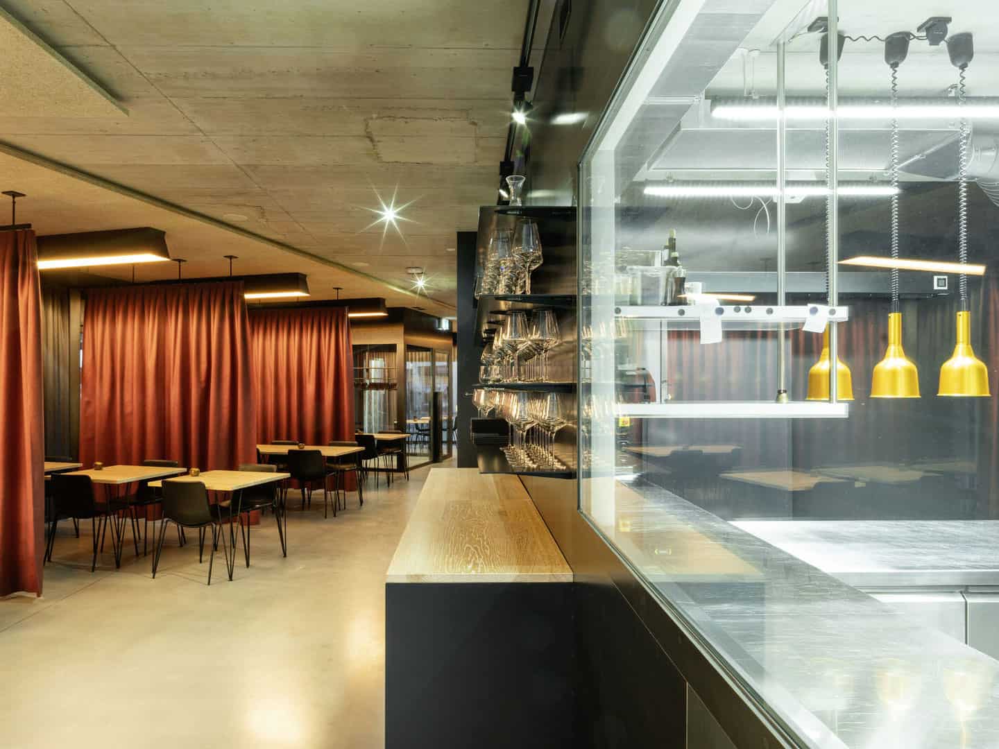 Bright modern restaurant interior with red curtains, wooden tables, black chairs, and an open kitchen with yellow pendant lights behind a glass partition.