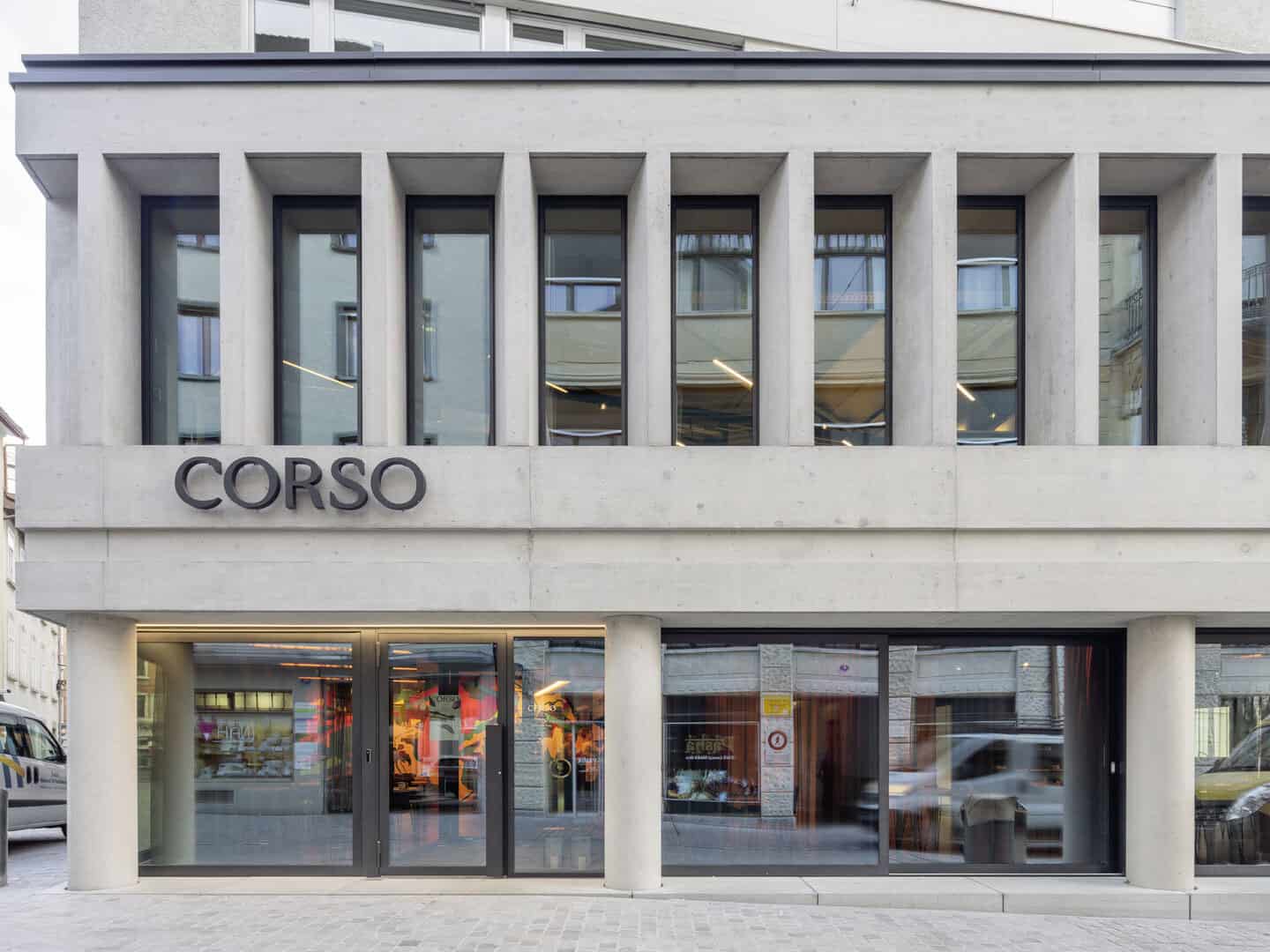 Contemporary building façade with large windows and the word "CORSO" displayed on the front, located in an urban setting with reflections of nearby structures visible in the glass.