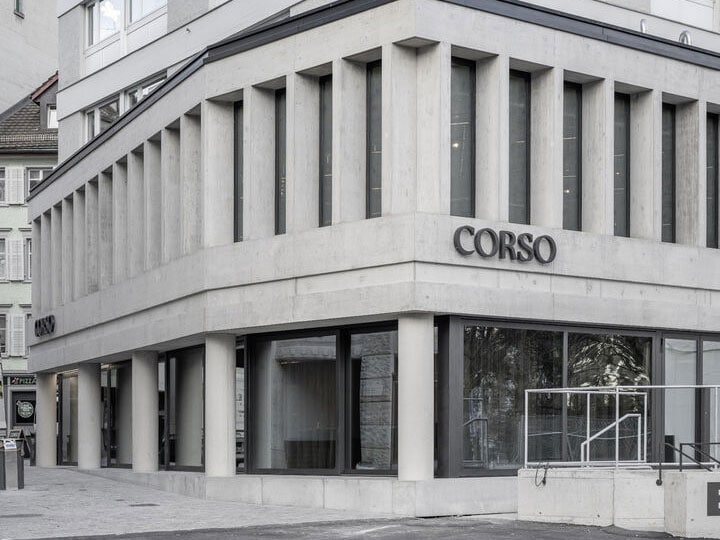 Modern corner building with concrete facade and large glass windows, featuring the sign "CORSO" on the exterior, situated in an urban area with neighboring structures.