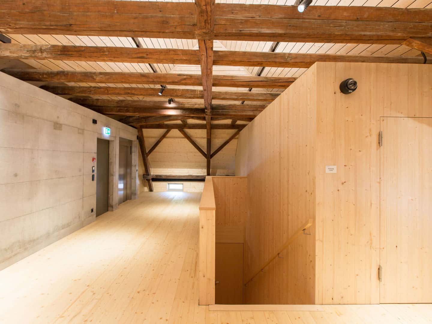 Polished wooden staircase in a modern building with exposed wooden beams on the ceiling. The space combines raw and finished wood elements for a contemporary, rustic aesthetic.