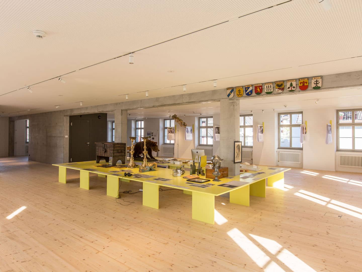 Colorful exhibit display with historical artifacts on a yellow table in a well-lit modern gallery space. The room features a wood floor, large windows, and walls decorated with coats of arms and artwork.