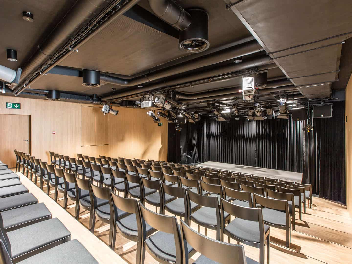 Empty auditorium with black curtains on stage and rows of chairs facing towards it, equipped with overhead lighting and audio equipment for performances or presentations.