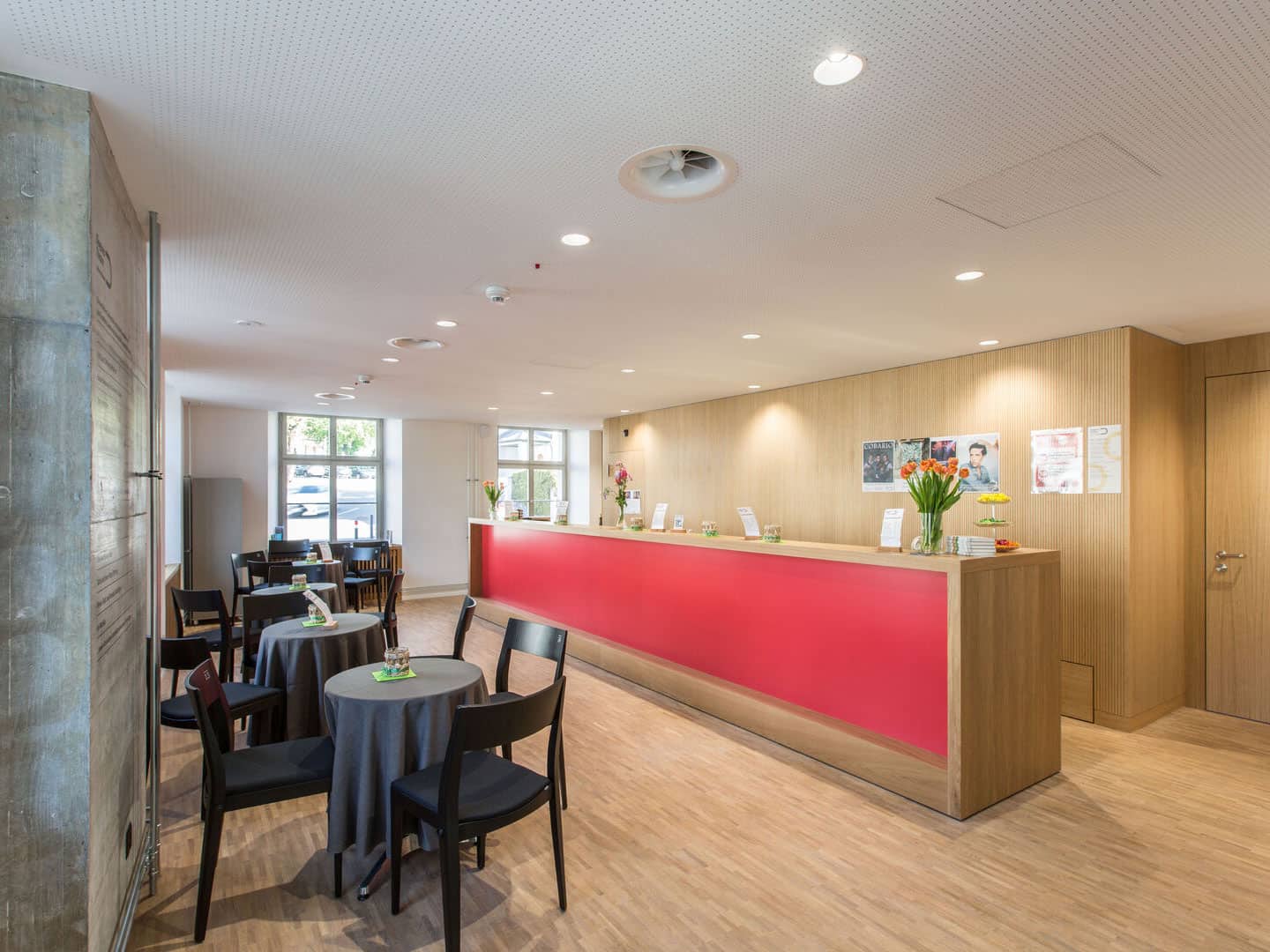 Bright and welcoming hotel reception area with a modern design, featuring a red accent counter, black chairs around tables, and colorful flower arrangements. Well-lit space with large windows.
