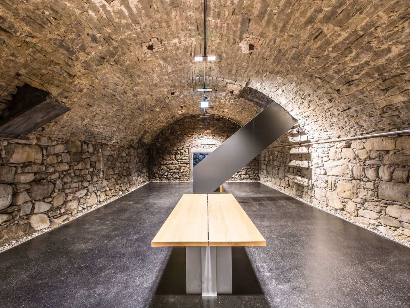 Weathered brick cellar with arched ceiling, modern gray sculpture, minimalist wooden table, and sleek lighting fixtures in an underground space.