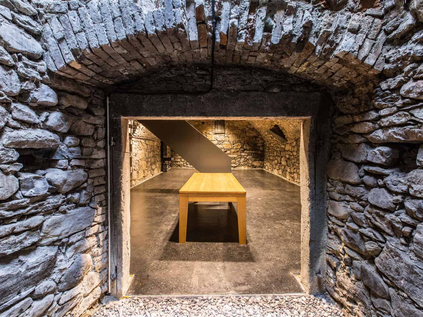 Rustic stone cellar interior with a wooden table, exposed brick walls, and a staircase, showcasing a historic underground vault used for storage or wine aging.
