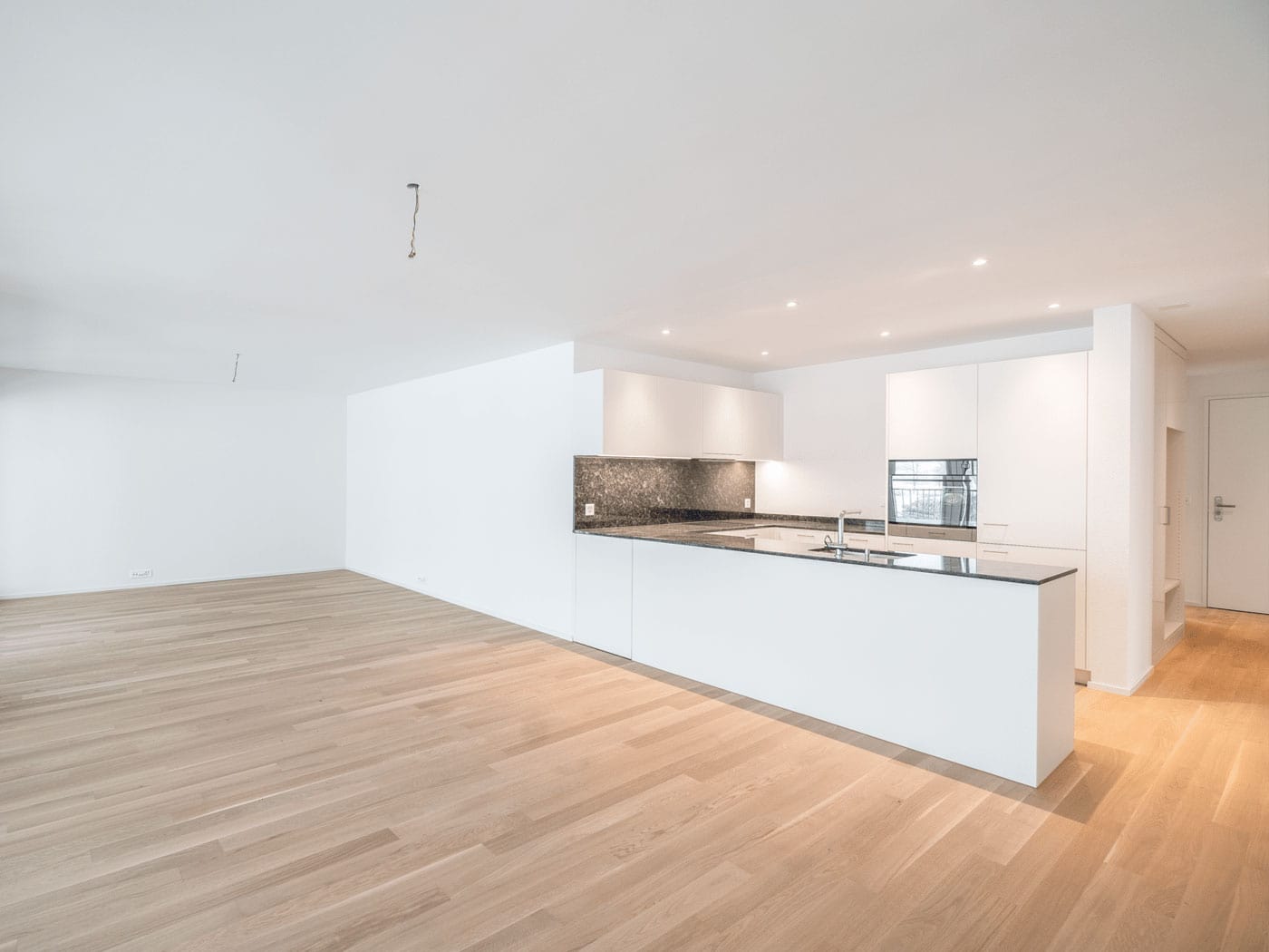 Bright, modern open-concept kitchen with minimalist white cabinets, black granite countertops, and integrated appliances. The space features sleek design elements, ample natural light, and light wood flooring, ideal for contemporary living.