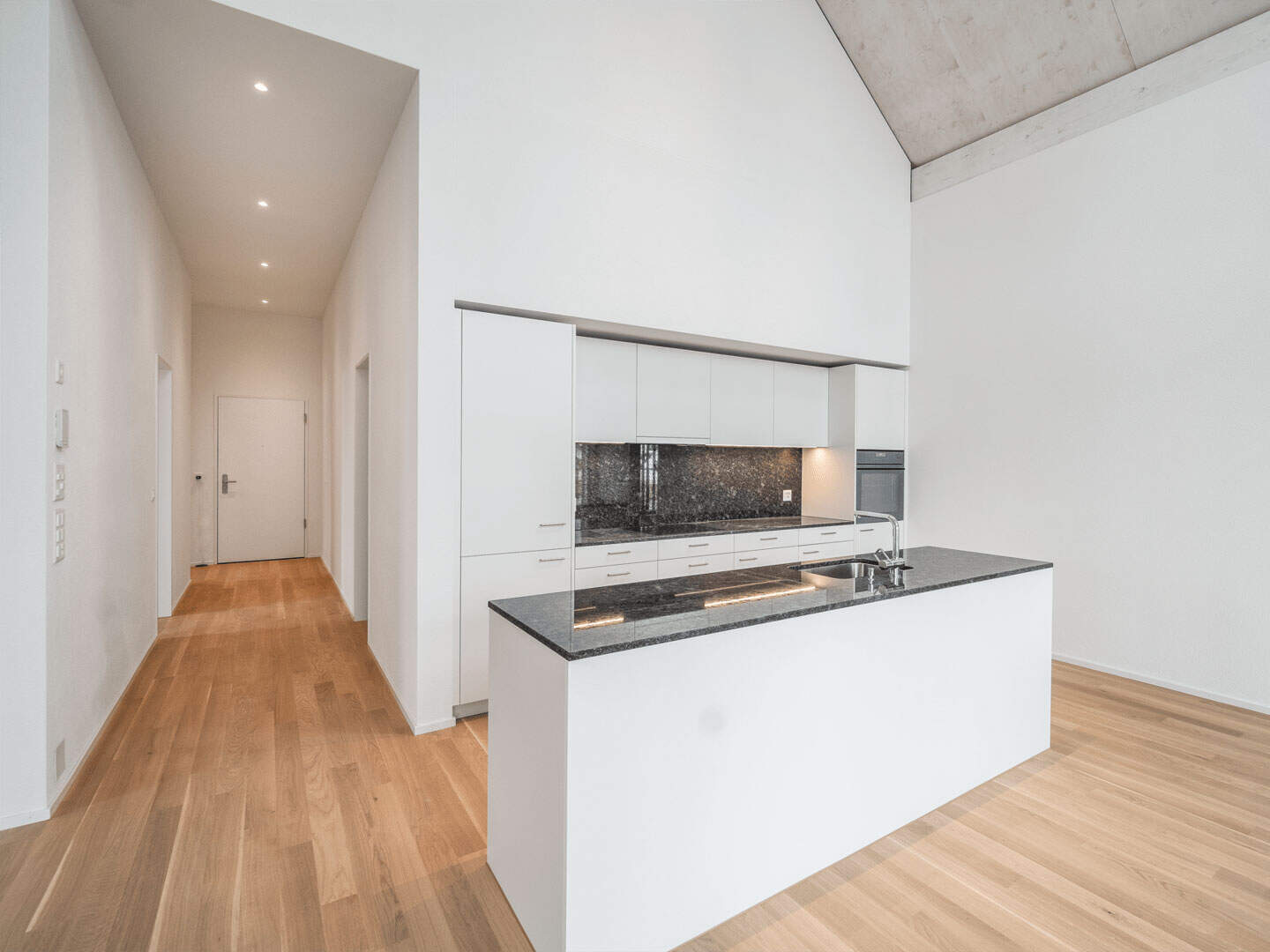 Modern minimalist white kitchen with black granite countertops, sleek cabinetry, and wooden flooring, featuring an open and airy design with high ceilings.