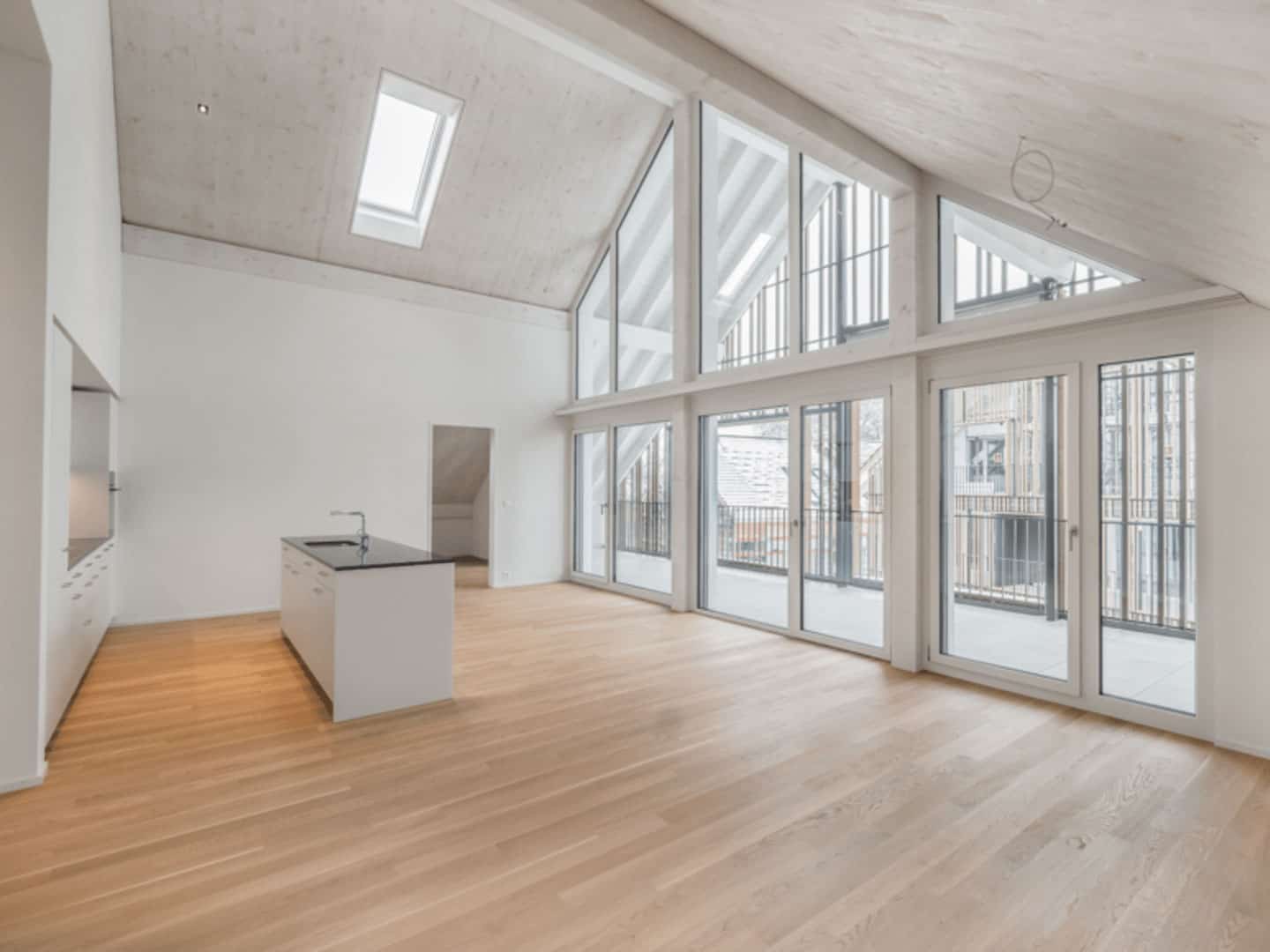 Bright, spacious modern living room with large floor-to-ceiling windows and skylights, featuring a minimalist kitchen area, high vaulted ceiling, and light wood flooring for a sleek, airy ambiance.