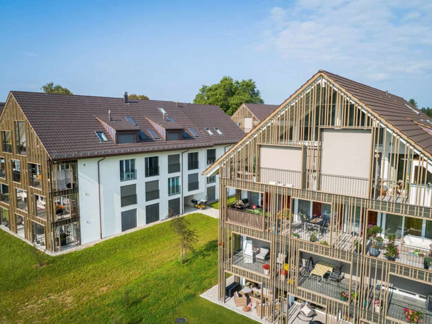 Modern multi-story residential buildings featuring wooden frameworks and spacious balconies, surrounded by green lawns and trees, with a bright blue sky in the background.