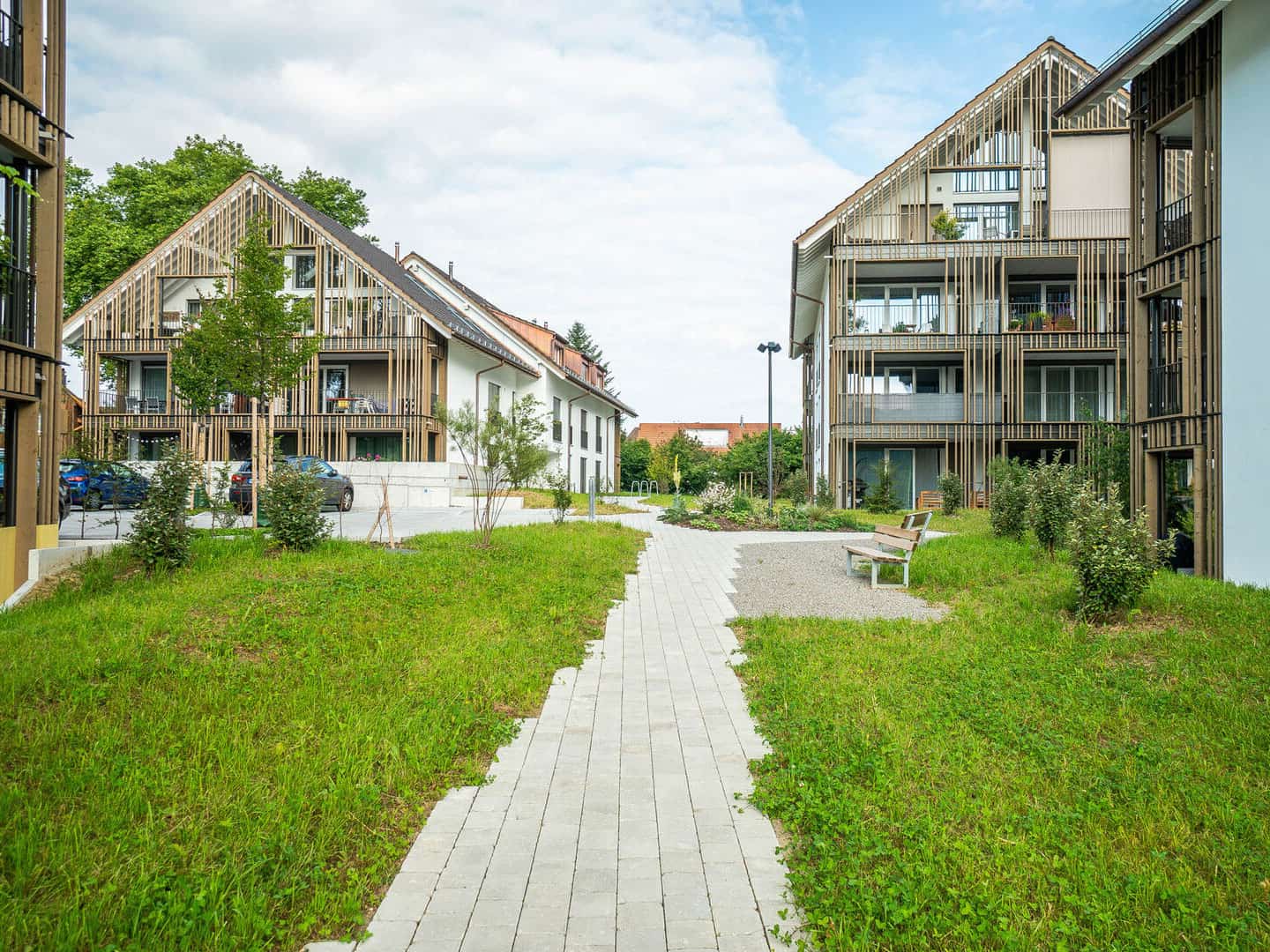 Modern residential buildings with wooden exterior accents and landscaped communal area featuring benches, small trees, and walking paths.