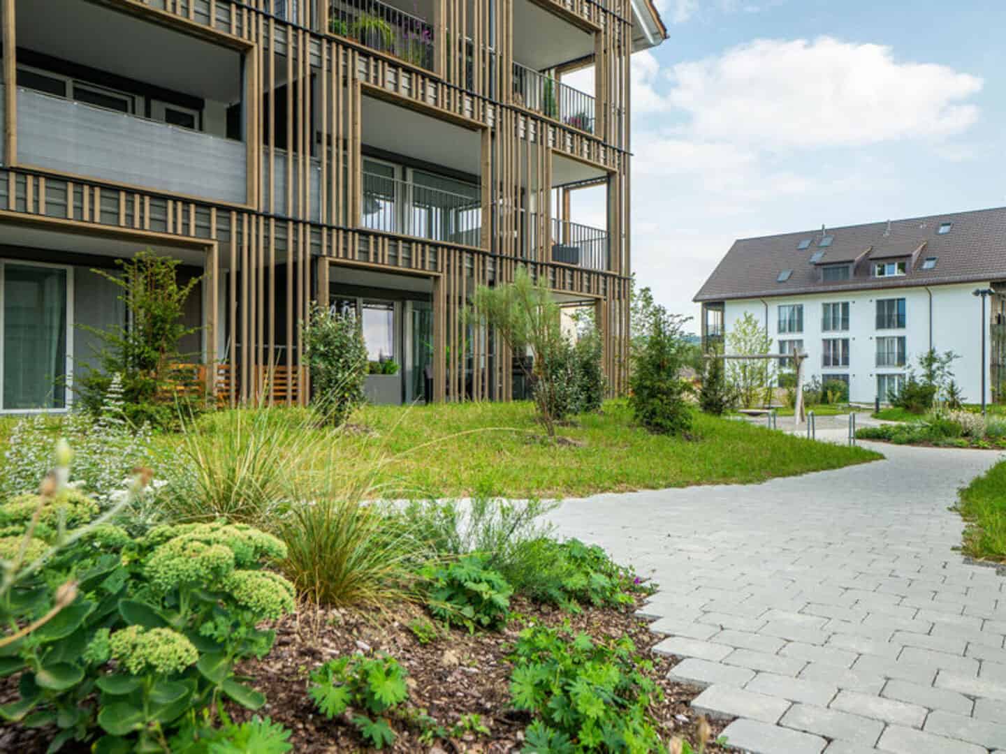 Lush green garden with landscaped plants and shrubs surrounding modern apartment buildings, paved pathway, and a clear sky in the background.