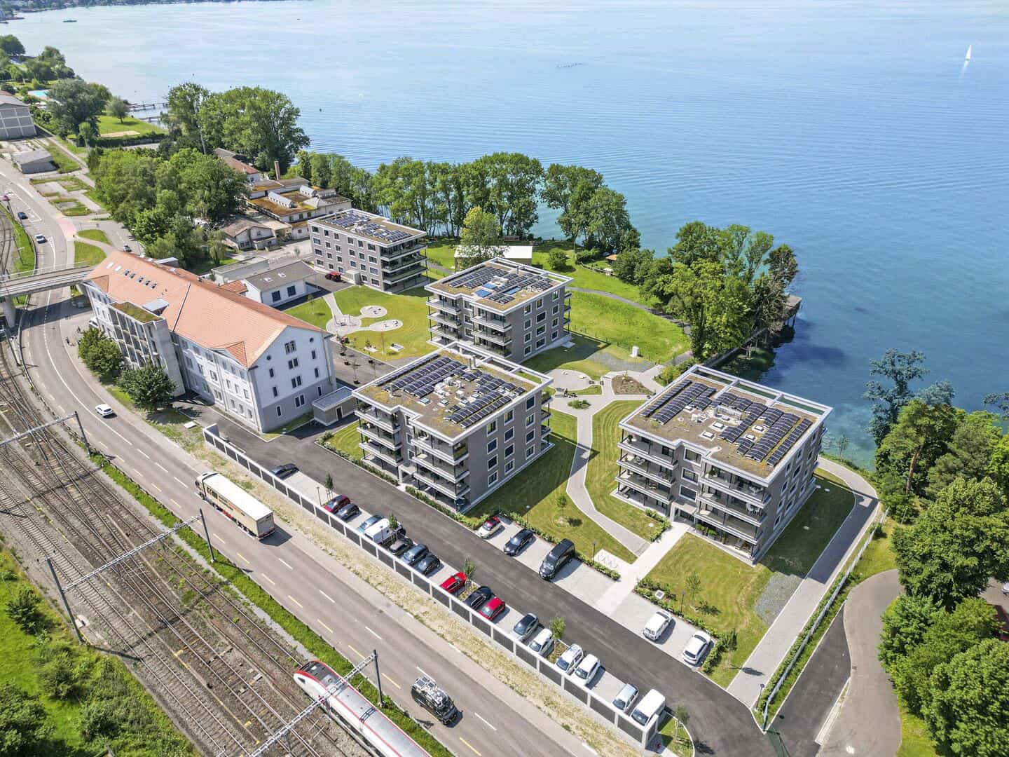 Modern lakeside apartment complex with solar panels and scenic water views, surrounded by greenery and located near railway tracks and a historic building.