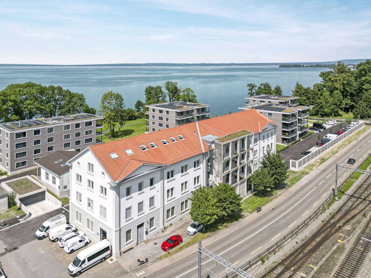 Modern residential buildings overlooking a large lake, featuring a mix of contemporary and classic architecture with greenery and parking in a scenic lakeside setting.