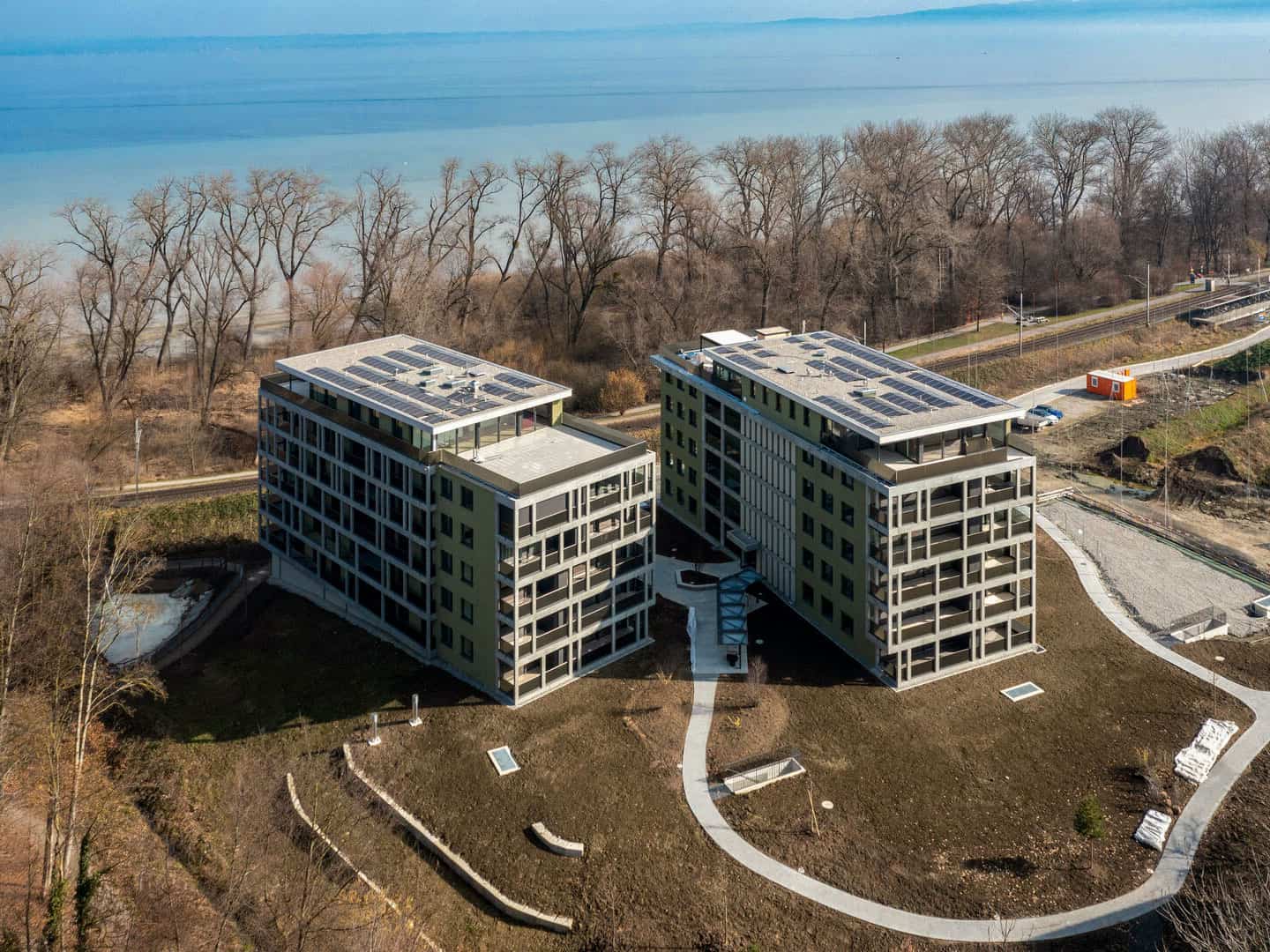 Modern two-story residential apartments with solar panels on the roofs, situated near a body of water and surrounded by trees and ongoing construction in a scenic setting.