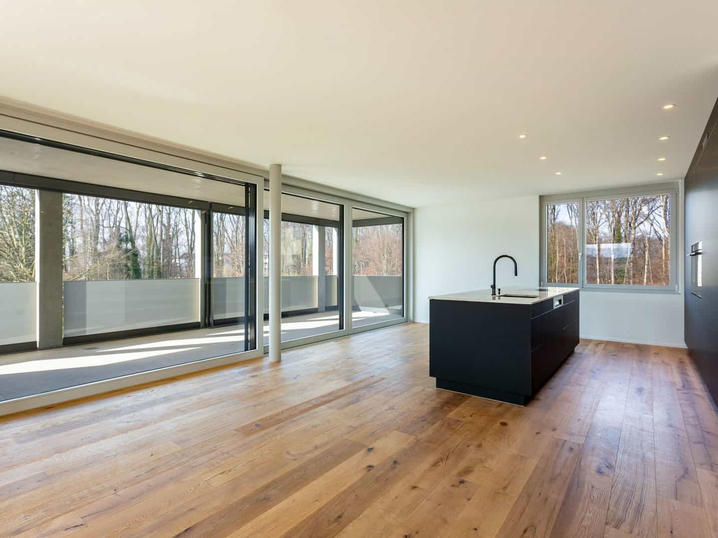 Bright, modern open-plan kitchen with large windows and sliding glass doors, natural wood flooring, and a sleek black kitchen island. The space connects to a balcony, offering scenic outdoor views and plenty of natural light.