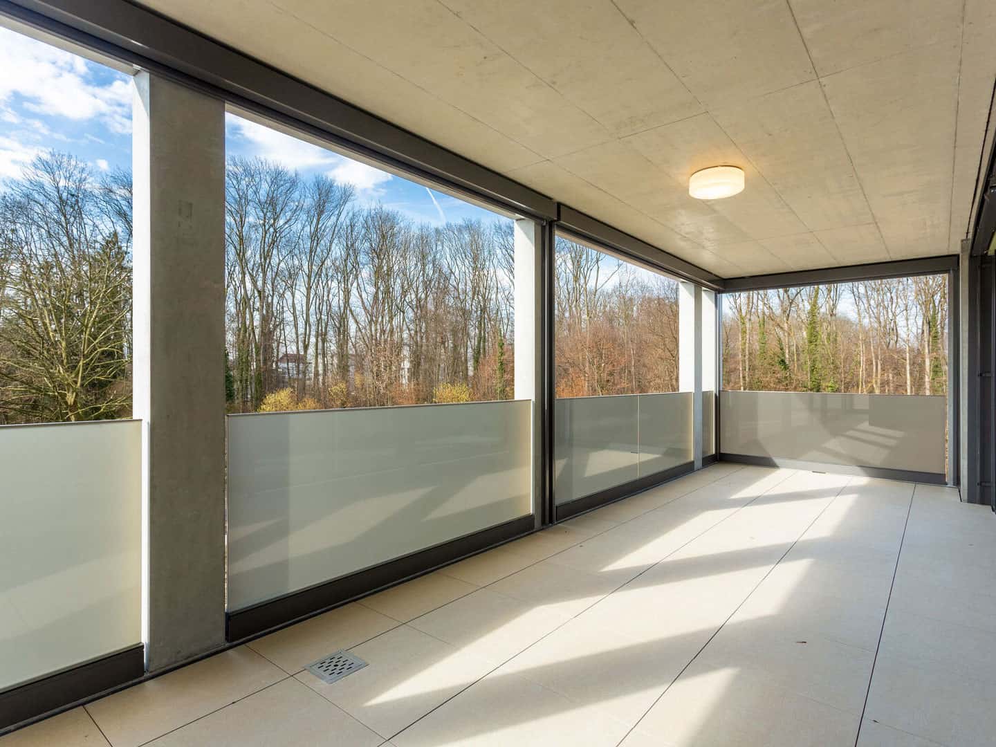 Bright enclosed balcony with frosted glass railings, overlooking a scenic view of leafless trees and a clear blue sky, ideal for relaxing or outdoor activities.