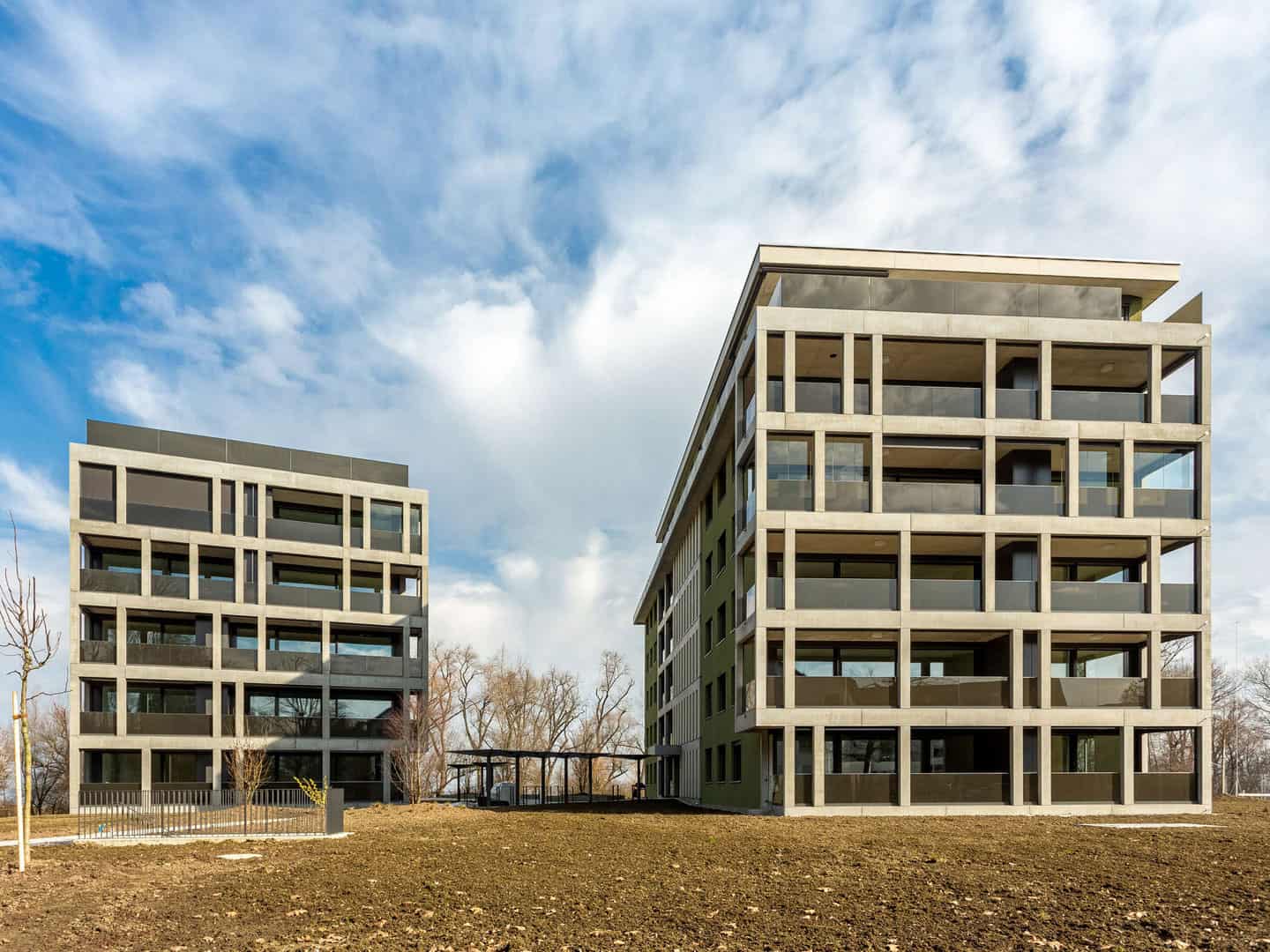 Modern residential apartment buildings with large glass windows and concrete frameworks, set against a partly cloudy sky in a suburban area.