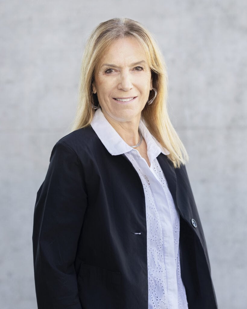 Elegant middle-aged woman with blonde hair, wearing a black blazer and white shirt, smiling confidently against a neutral background.