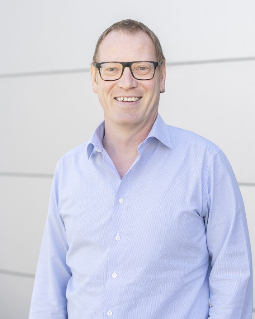 Bright smiling man wearing black glasses and a light blue dress shirt, standing outdoors against a modern white wall with horizontal paneling.