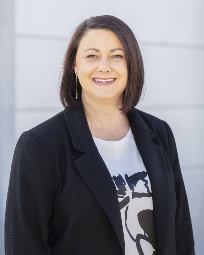 Smiling professional woman with shoulder-length dark hair, wearing a black blazer and graphic t-shirt, outdoors against a neutral background.